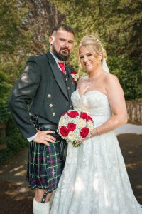 Bride holding a red and white bouquet beside a groom in a tartan kilt at Webbington Hotel